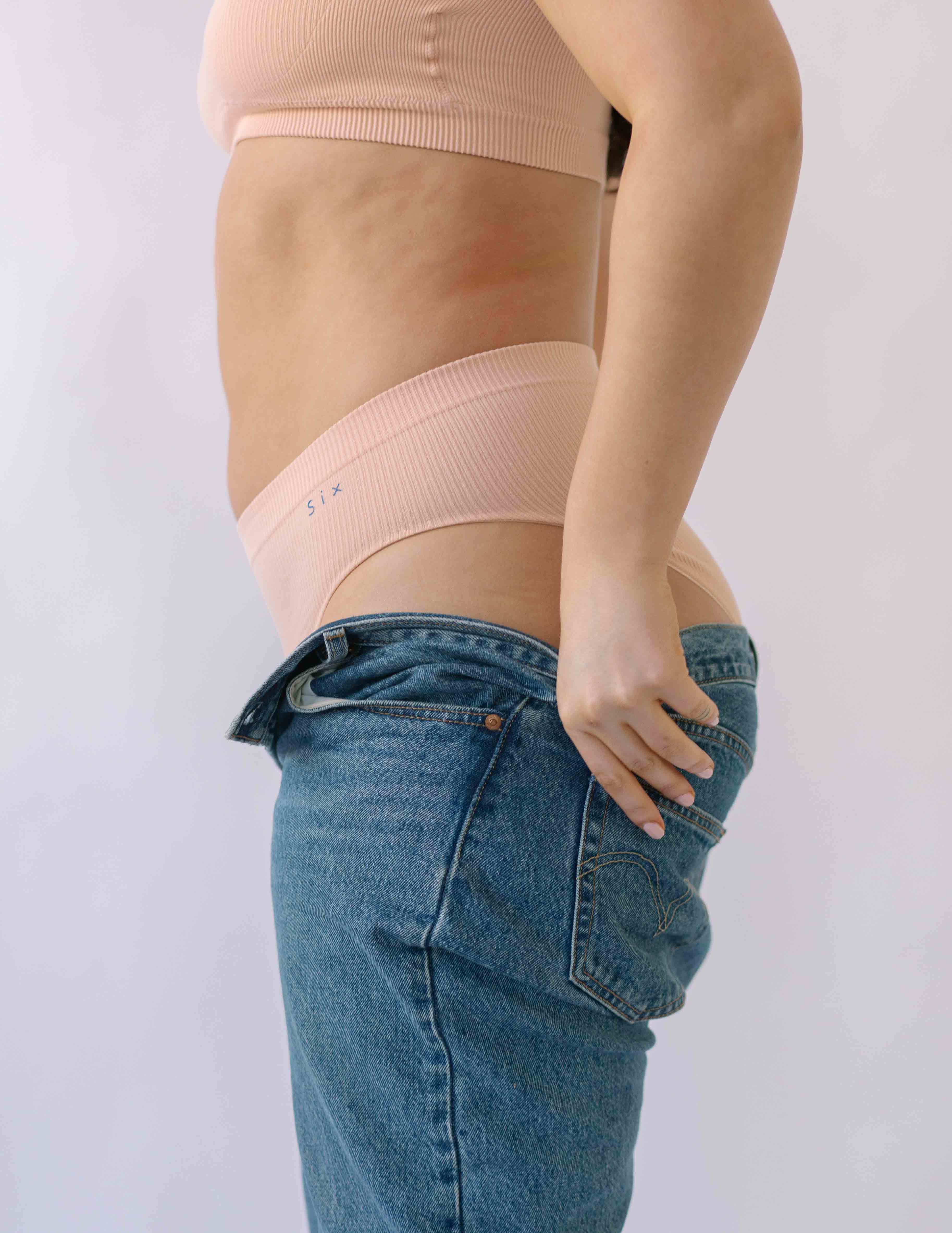 Woman putting on a pair of jeans  wearing the Ada ribbed seamless high waisted knicker from Six. Suitable to wear throughout pregnancy, maternity and as a supportive post C section knicker.