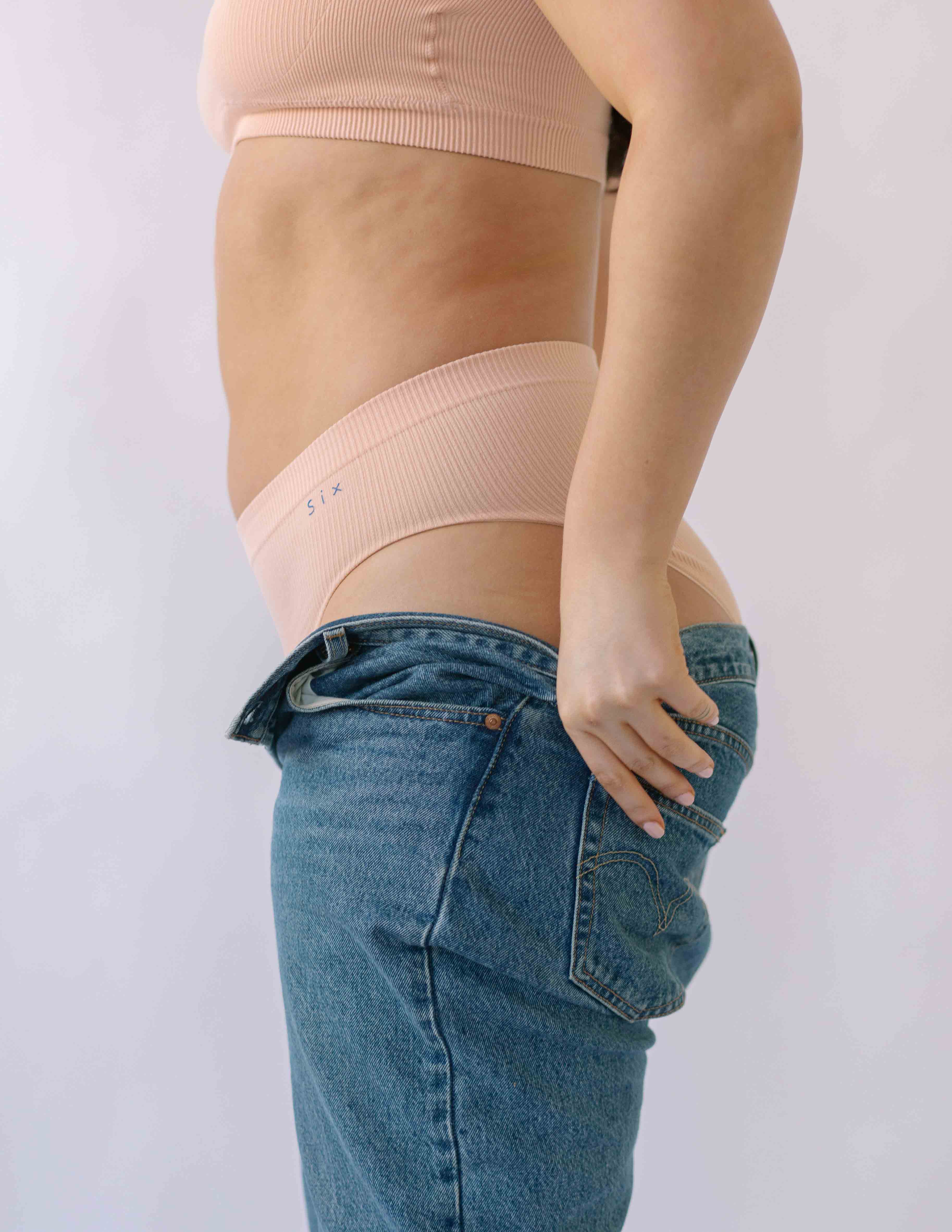 Woman putting on a pair of jeans wearing the Ada ribbed seamless high waisted knicker from Six. Suitable to wear throughout pregnancy, maternity and as a supportive post C section knicker.