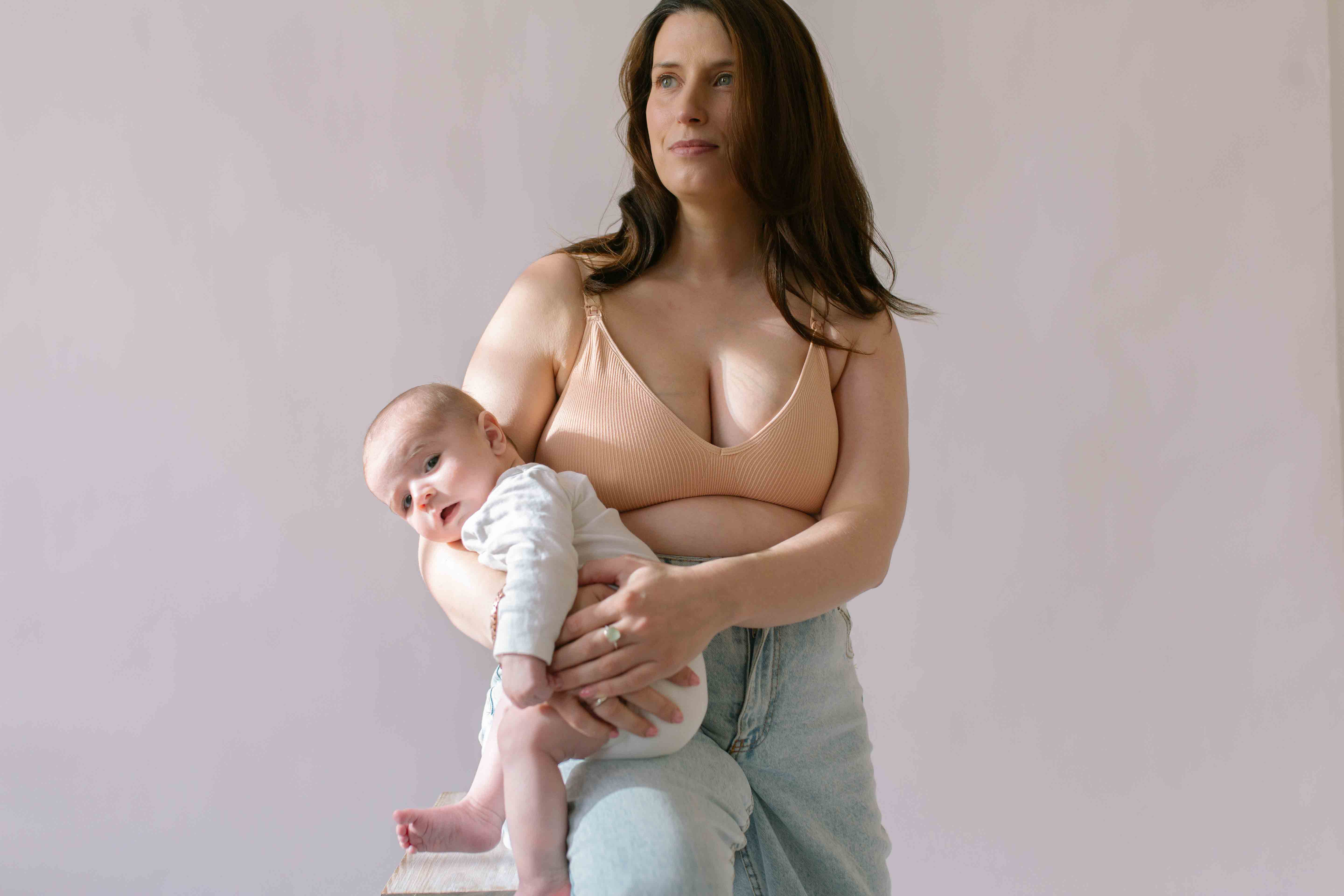 Woman sat holding her baby wearing the Six Ada seamless ribbed nursing bra in Nude Blush colourway. Comfortable and supportive nursing bra to be worn through pregnancy and breastfeeding.